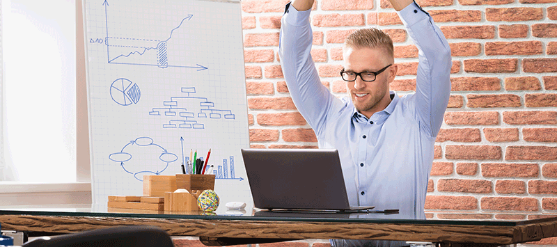 Person in Businesskleidung streckt die Arme über den Kopf, während sie auf einem Fitnessball an einem Schreibtisch mit Laptop sitzt, was auf einen Online-Firmenfitnesskurs hinweist. Im Hintergrund zeigt ein Whiteboard verschiedene Diagramme und Grafiken, die den Fokus auf Produktivität und Gesundheitsmetriken legen.